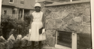 Old black and white image of Monica Ross dressed in white church dress, white gloves, white hat, and holding a black clutch