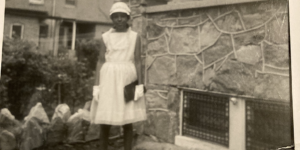 Old black and white image of Monica Ross dressed in white church dress, white gloves, white hat, and holding a black clutch