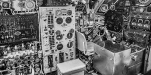 Black and white image of an incredibly complicated panel and seat in the pilot's part of the airplane. The image is a scene of extreme complexity with an empty pilot's seat on the right and dials and knobs throughout the image.
